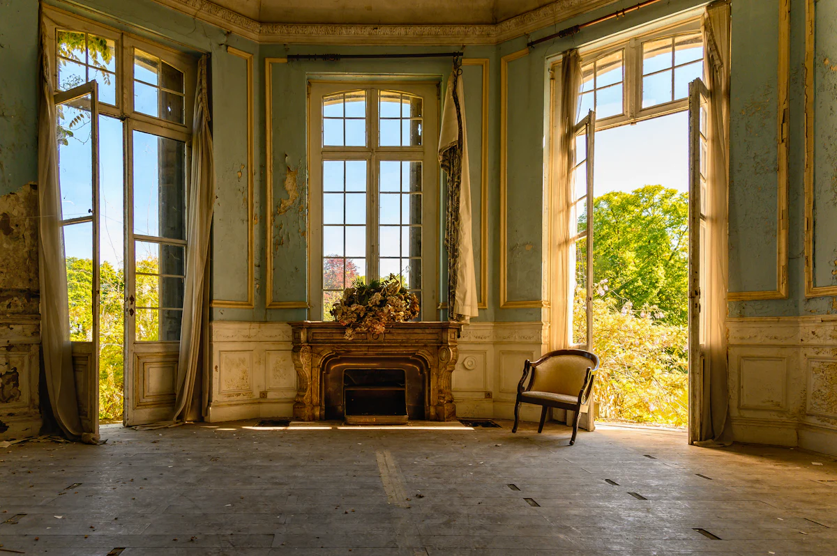 Grande pièce ancienne vidée, baignée de lumière, avec une cheminée et un fauteuil
